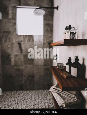 Bathroom wooden shelves with bath utensils and cosmetics in modern gray bathroom with shower and pebbles on floor, contemporary Stock Photo