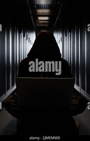 The silhouette of the hacker in a hood, the burglar sits with the computer among servers in the dark. Storage of data, cloudy se Stock Photo