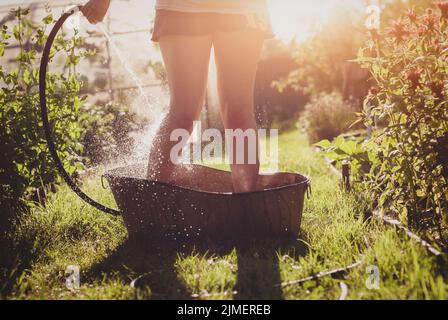 Bathing in the summer garden - woman pours cold water on her feet from garden hose standing in an iron bath on garden path in su Stock Photo