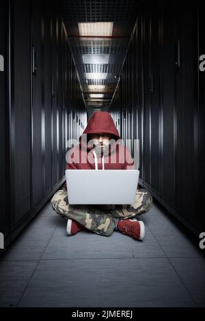The hacker in a hood sits with the computer among servers, the burglar. Storage of data, cloudy services, system breaking, attac Stock Photo