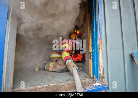 A firefighter wearing breathing apparatus feeds hose from the doorway to fellow firefighters farther in the building as members of the East Hampton Fi Stock Photo
