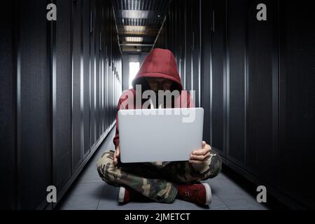 The hacker in a hood sits with the computer among servers, the burglar, face covered with a hood. Storage of data, cloudy servic Stock Photo