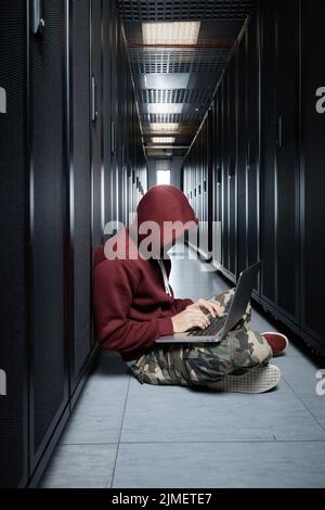 The hacker in a hood sits with the computer among servers, the burglar, face covered with a hood. Storage of data, cloudy servic Stock Photo