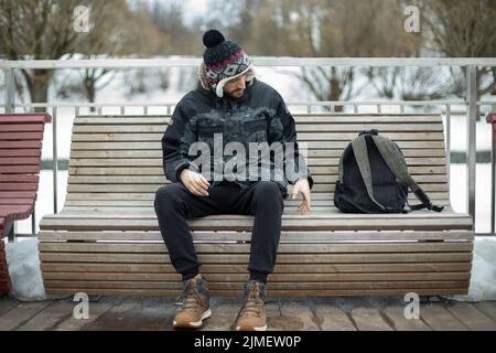 Guy sits on bench in park. Guy with backpack on street. Man in warm hat. Student rests. Stock Photo