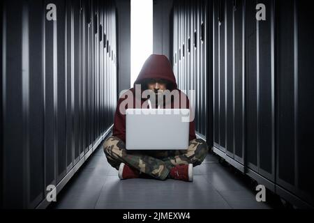 The hacker in a hood sits with the computer among servers, the burglar, face covered with a hood. Storage of data, cloudy servic Stock Photo