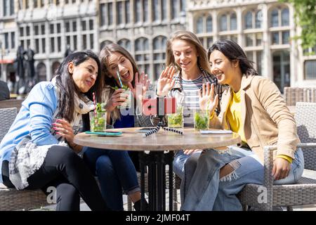 Four multi ethnic female friends, tourists or students sitting at a cafe terrace in the city center using mobile phone for a vid Stock Photo
