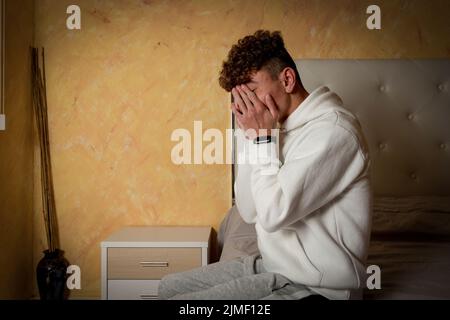 Stressed, depressed or grieving man sitting on bed covering face with hands and crying at home. Worried and frustrated male suffering depression or Stock Photo