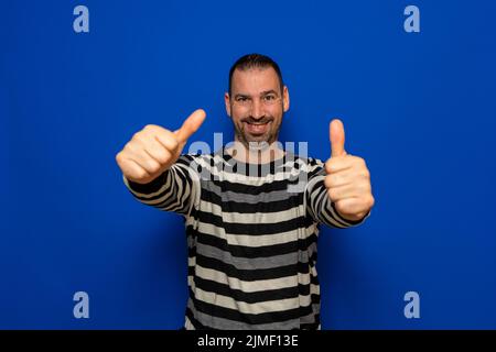 Attractive cheerful fit bearded man in striped pullover standing isolated over blue background, showing thumbs up. Stock Photo