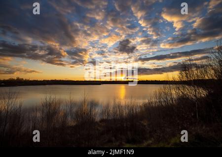 A beautiful blue lake with some tree silhouettes in the foreground under a dramatic sunset sky Stock Photo