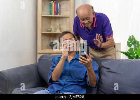 Asian senior holding mobile smart phone communication online internet video call together on sofa at home. Stock Photo