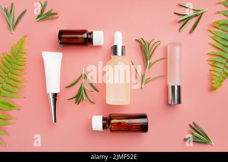 Glass and plastic cosmetic bottles on pink background with fern and rosemary leaves Stock Photo
