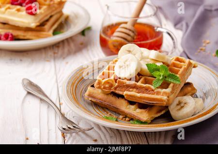 Belgian waffles served with banana and mint leaf on white wooden kitchen table with syrup aside Stock Photo