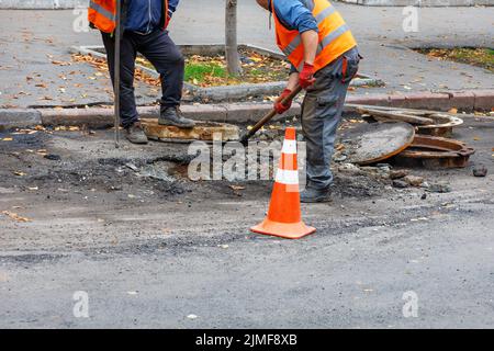 A team of road workers with a shovel and a crowbar clean a place for a new sewer manhole on the roadway on a summer day. Copy space. Stock Photo