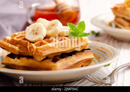 Belgian waffles served with banana and mint leaf on white wooden kitchen table with syrup aside Stock Photo