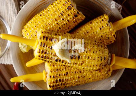 Top view of Wooden table with deep grilled sweet corn cobs under melting butter with plastic holder on clay dish Stock Photo