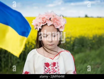 Close-up portrait of sad Ukrainian girl 7 years old in traditional embroidered blouse with blue-yellow flag on background of yellow blooming rapeseed Stock Photo