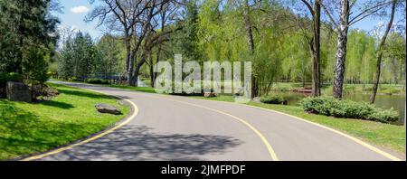 Road through the park. Asphalted path for pedestrians and cyclists through beautiful greenery and a lake in the park. Stock Photo