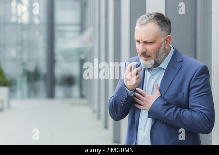 Senior male businessman sick near office, boss uses inhaler to ease breathing, gray haired banker has asthma attack Stock Photo