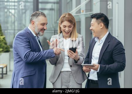 Diverse business team, male and female business people together outside office, group of successful business people looking at colleague's phone, smiling and happy Stock Photo