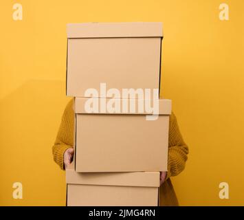 A woman in a sweater holds a stack of brown cardboard boxes on a yellow background. Relocation Stock Photo