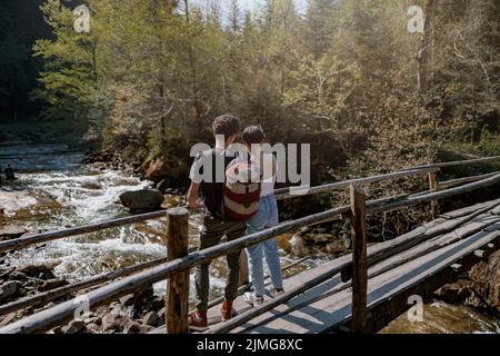 Back view on couple of young hikers standing on wooden bridge over fast stream and hugging. Stock Photo
