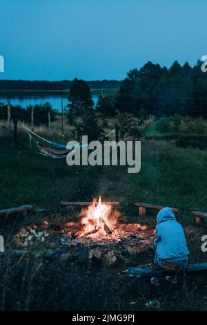 A person with a hood sitting next to the bonfire with hammocks, forest and lake in the background. Stock Photo