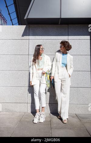Two woman friends have a rest after walking in city standing near wall Stock Photo