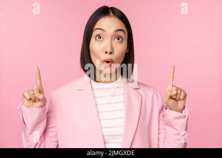 Enthusiastic corporate worker, asian business woman pointing fingers up and smiling, showing advertisement, logo, standing over Stock Photo