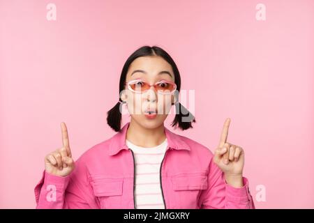 Close up portrait of asian woman gasping, looking surprised, pointing finger at banner, advertisement, standing over pink backgr Stock Photo