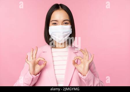 Asian businesswoman in suit and medical face mask, showing okay sign, recommending using protective equipment in office during c Stock Photo