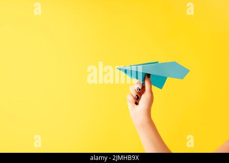 A woman's hand holds blue paper airplane on yellow background Stock Photo