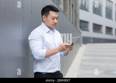 Thinking asian businessman reading news using tablet computer outside office building, man focused and serious holding electronic device Stock Photo