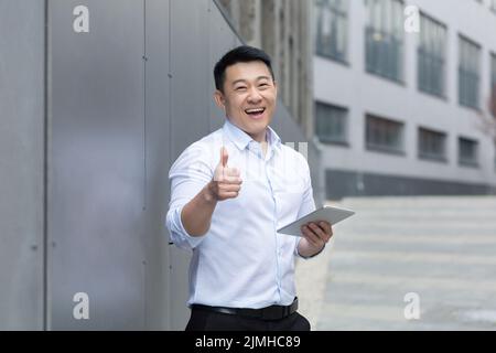 Happy and successful businessman asian looking at camera and thumbs up, man with tablet computer standing outside office building Stock Photo
