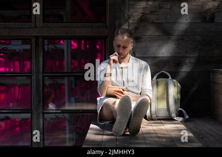 Young man sits on wooden stairs outdoors writing notes in paper pad in rays of sunlight Stock Photo