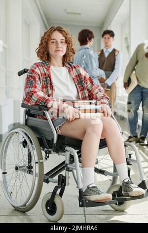 Cute teenage female student in wheelchair sitting in front of camera in college corridor against group of her classmates talking at break Stock Photo