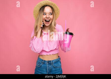 Photo of beautiful positive emotional young blonde woman wearing pink crop blouse and straw hat talking on cellphone while drink Stock Photo