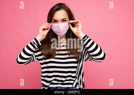 Photo shot of young beautiful brunette woman in reusable virus protective mask on face against coronavirus isolated on the pink Stock Photo