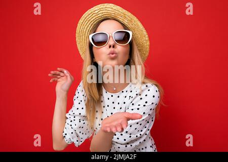 Photo shot of young beautiful cute happy blonde woman wearing casual dress, sunglasses and straw hat isolated over red backgroun Stock Photo