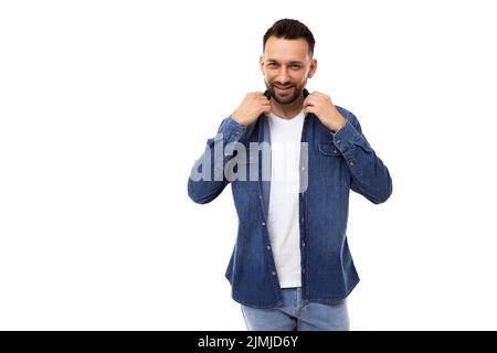Stylish elegant man in a white t-shirt and denim light blue shirt straightens clothes by the collar smiling looks at the camera Stock Photo