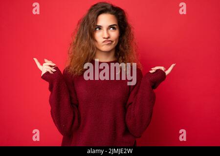 Photo of young positive thoughtful beautiful brunette curly woman with sincere emotions wearing casual red pullover isolated on Stock Photo