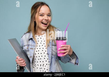 Image of joyful european blond woman smiling and standing against blue wall isolated with silver notebook and takeaway coffee in Stock Photo