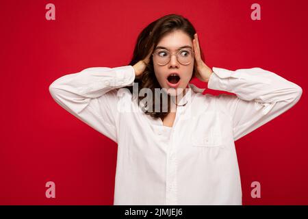 Beautiful young shocked amazed curly brunette woman wearing white shirt and optical glasses isolated on red background with copy Stock Photo