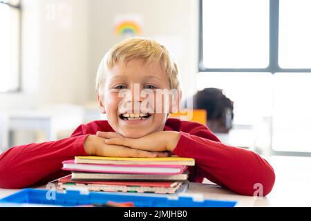 Portrait of smiling caucasian elementary schoolboy leaning on stacked books at desk in classroom Stock Photo