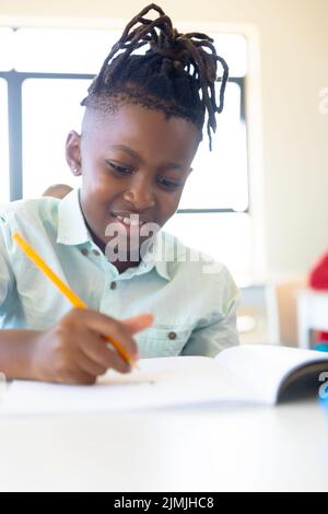 Smiling african american elementary schoolboy writing on book while sitting at desk in school Stock Photo