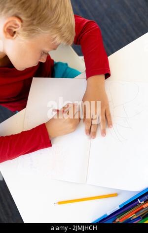 High angle view of caucasian elementary schoolboy writing on book while sitting at desk in school Stock Photo
