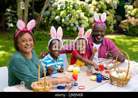 Portrait of happy african american siblings and grandparents in bunny ears painting easter eggs Stock Photo