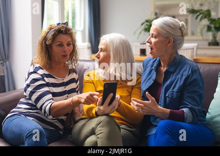 Surprised multiracial senior female friends looking at smart phone while sitting on sofa at home Stock Photo
