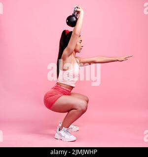 Fully focused on her form. Studio shot of a sporty young woman doing squats with a kettlebell against a pink background. Stock Photo