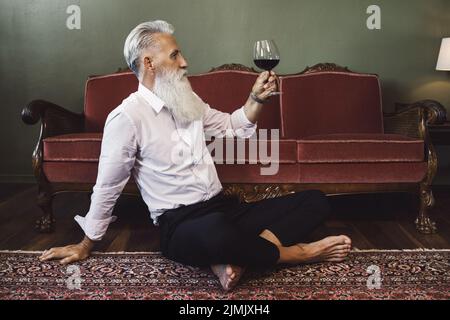 Handsome bearded senior man sitting on the floor and drinking red wine Stock Photo