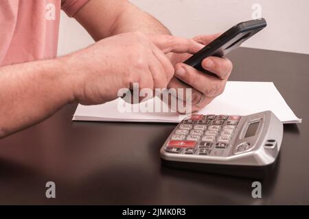 Unrecognizable Latino adult male checking messages on his cell phone while doing math with calculator. Concept of business, investments, communication Stock Photo